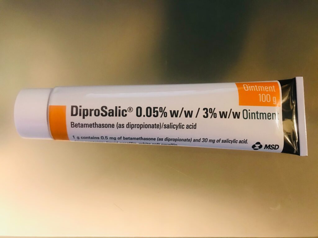 Kleine Salbentube mit weißer Creme auf einer hellen Fläche, daneben der geöffnete Deckel